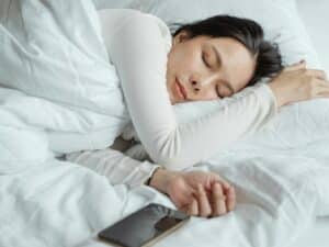 Calm Asian female wearing white pajama sleeping in comfortable bed with white sheets near modern mobile phone in morning